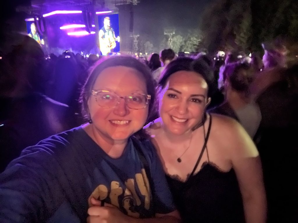 Kelley Sanabria and a friend smiling in the crowd at a Zach Bryan concert, with Zach Bryan on the big screen behind them